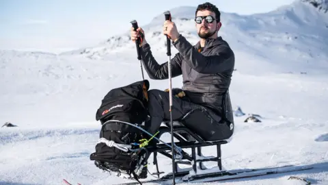 Aman sits on a sled with skis attached to the bottom while he holds two ski poles. He is in the middle of a snowfield with snowy hills in the background.