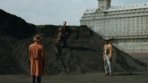 A24 A scene from The Brutalist featuring three men. One man, who has dark hair and clothes, is stood on a large pile of rubble. In front of him are two other men, one in a brown hat and coat with his back to the camera, and the other in a casual brown jacket and blue jeans and flat cap. Behind them is a grand grey building with the letters MAH on its roof.