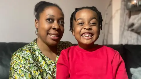 A Mum in a green flowery top cuddles her young son who is wearing a bright red top. Both have beaming smiles.