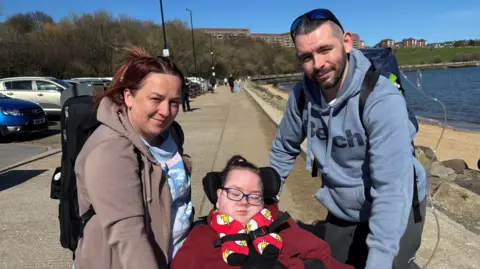 Stephen and Laura Davidson are on the pavement next to the sea front with Milo in a wheelchair. To the right you can see the sea, sand and coastline. The woman and man both have oxygen canisters on their backs. They are looking at the camera and smiling while holding the boy's hand.