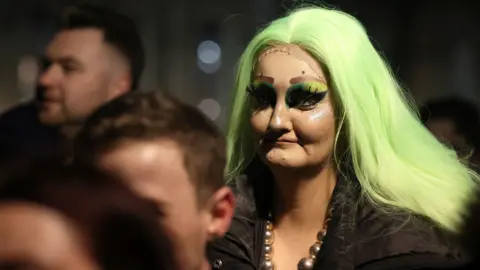 Reuters A person in a bright green wig with strong eye make up looks away to the left of camera, with other blurred faces around her