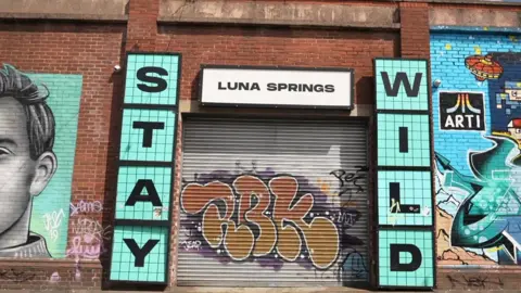 A grey metal shutter with orange graffiti is pulled over the entrance to Luna Springs. Murals are painted on the red brick wall around its entrance. Signs either side of the entrance say 'Stay Wild' in blue, with a white 'Luna Springs' sign above.