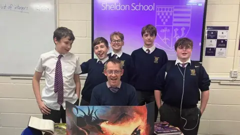 A group of students gathered around a dungeons and dragons board game. The children are standing in front of their teacher, while a blue screen reads "Sheldon school" on the wall behind them