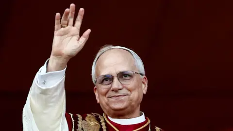 Reuters Pope Leo, wearing glasses and religious dress, waving against a red background