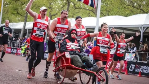 RFU IPF Gareth Rees in his specially adapted wheelchair and his team of runners pushing him at the London Marathon in 2024.  They are all wearing red and white IPF charity bids with other runners behind and supporters lining the street.