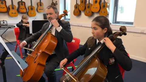 In the forground on the right is Lucy, a young school girl wearing a school uniform, looking at sheet music on a stand in front of her while she plays the cello which is leaning on her left shoulder. Next to her is Mr Perre, a teacher also playing the cello on his left shoulder while looking at sheet music. He's wearing a suit and piano tie.In the background is two windows, and the wall is covered in acoustic guitars hanging up.