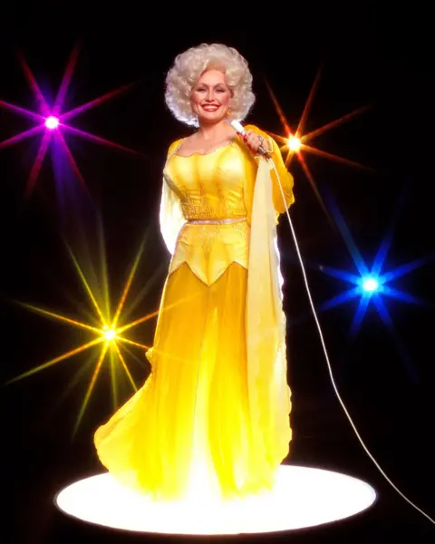 Harry Langdon/Getty Images Dolly Parton poses for a portrait session in 1978. She holds a microphone against a black backdrop and wears a bright yellow flowing outfit. 