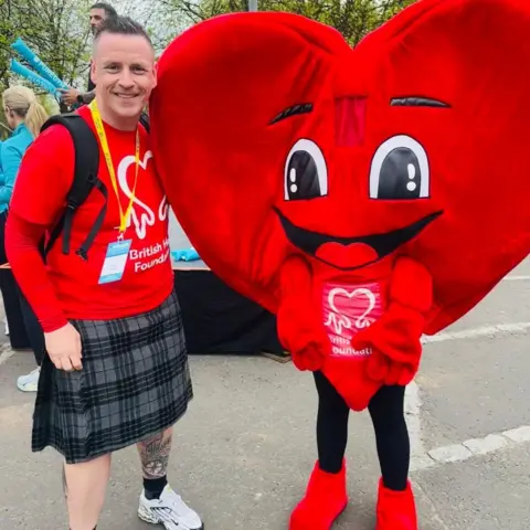PA Media Neil Magill is standing next to someone dressed as a red heart with a smiling face. They are both wearing red British Heart Foundation T-shirts, Neil is in a grey tartan kilt. 