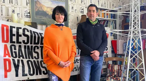 BBC Eileen West and Justin Rowlatt pictured in her study, which has wallpaper which looks like bookshelves. She has straight, dark hair cut into a bob with a fringe. She wears a bright orange asymmetric tunic with long sleeves, and patterned leggings. She is clasping her hands in front of herself. Justin has short dark hair, jeans and a dark fleece. Behind them is a big banner which reads 'Deeside against pylons' and next to Justin is a model of a pylon, which is taller than him. 