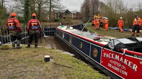 dark blue and red canal boat in a small section of canal blocked off with metal dams. it is in water and there are dozens of people standing on banks either side of it in orange outfits