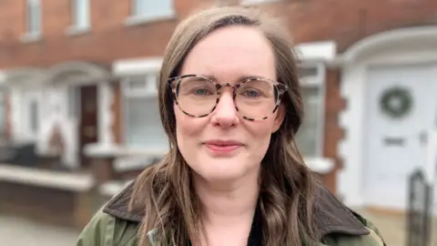 A woman wears clear spectacles as she smiles to the camera. She has long brown hair and wears a dark-green waterproof coat. Behind her, out-of-focus, is a row of two-storey red-brick houses. 