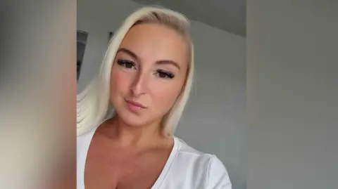 Jody Shovelton Jody Shovelton - a blonde woman, brown eyes, false lashes, white t-shirt, selfie. Jody