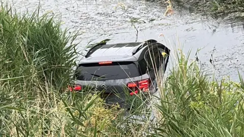 A silver and black car has entered a river which has several bits of grass floating on it. The river bank is on the left side of the image.