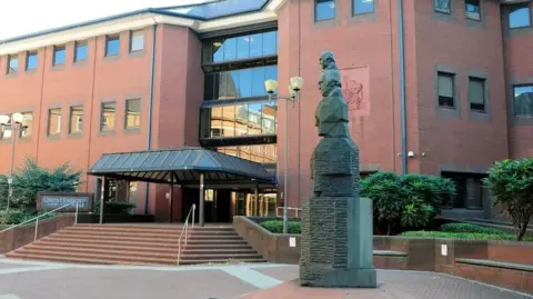 Looking to the entrance of a large red brick building, which has a number of steps up to the wide glass front doors. There is a sculpture in the foreground showing a series of busts stacked on top of one another.