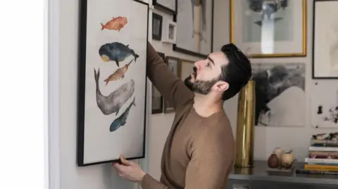 Getty Images Young man wearing a brown jumper hangs a painting on a wall 