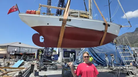 Rob Donald A big 9.8 metre boat has been lifted out of the dock in Cape Town by a crane and massive strops. A girl in a pink t-shirt and wearing sun glasses is doing a thumbs up in front of it. The scene is busy and industrial. 