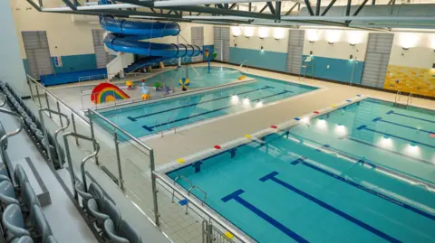 An aerial view of Newry Leisure Centre's pool hall taken from its viewing gallery.  There is a 25m main pool, a smaller learner pool and a children's play area.  A large blue flume curls down from just below the ceiling. 