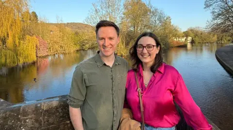Laura-May Nardella Laura-May Nardella and her husband Ryan Monk are smiling at the camera, with a lake and trees behind them. Laura-May is wearing a bright pink shirt with glasses and has brown shoulder-length hair. Ryan is wearing a great shirt and has short brown hair