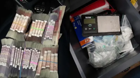 Bundles of money on a car seat, alongside small plastic bags in a pile, a set of black digital scales electro and what appears to be a blue packet of tablets.