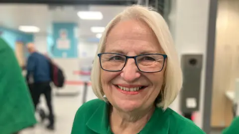 Elizabeth Baines / BBC A woman with white blonde hair wears glasses and smiles. She wears a green polo shirt