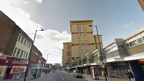 Street View image showing a road with shops on either side and tower blocks up ahead.