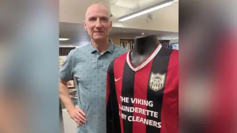 LDRS Gary Maguire wearing a grey top with his hand on his hip standing with Frank Sidebottom's black and red striped Big Shorts football shirt which is in the Frank's World Exhibition.