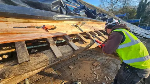 Historic Property Restoration Ltd A workman in a yellow high-vis vest and hard hat works on the rafters