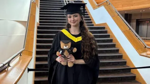 Oksana Lohvynenko A brunette girl stands in her black cap and gown, that features a yellow satin hood sat draped around her front. She holds a brown teddy bear in her both hands, who also wears a miniature cap and gown. The girl is stood in front of a set of stairs.