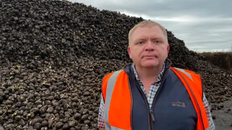 A blonde man in a high visibility jacket stands in front of a huge pile of sugar beet that has been farmed from his land