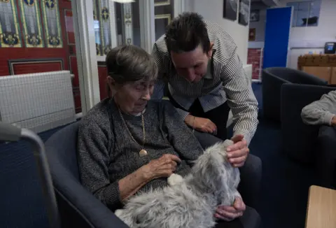 Facundo Arrizabalaga / MyLondon Female care home resident holds toy dog (left) while a man (right) pets a toy dog held
