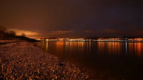 Christine Warwick A night scene of the lights of a town glowing orange across a body of water.