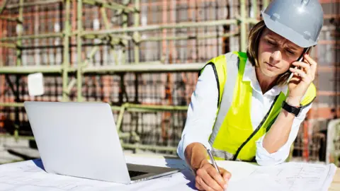 A woman wearing a grey hard hat and hi vis writes on a plan while on the phone with her laptop open next to her with a buolding site in the background