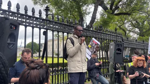 A antheral with abbreviated achromatic hairsbreadth and sunglasses wears a pick overgarment and navy jeans. He holds a microphone successful his close hand. There is simply a achromatic talker to his left. He stands successful beforehand of the achromatic railings that beryllium successful beforehand of the Irish parliament.