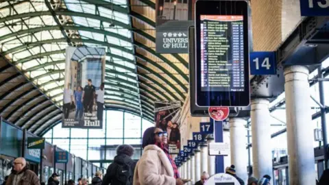 Hull Travel A busy Hull Paragon Interchange bus station. The focus is on a woman wearing black sunglasses and a thick beige coat looking at a digital board showing a list of bus times.