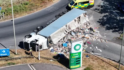 A white lorry is on its side next to a roundabout. What appears to be mounds of rubbish is coming out of the lorry. In the foreground is a white and green BP advertising pillar. Behind the lorry is a yellow and green ambulance parked in the road