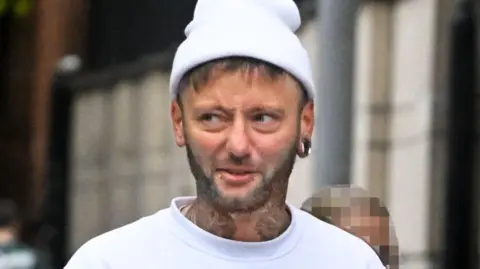 A man with a short beard, neck tattoos, two piercings below his bottom lip and a large circle earring in his ear wears a white beanie. He also wears a white sweatshirt as he walks through a street. 