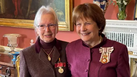Suffolk County Council Lady Clare Fitzroy, Countess of Euston, wearing a burgundy coat and a pearl necklace while standing next to a woman wearing a brown coat over a burgundy top and a gold necklace and a medal. They are both smiling and looking past the camera.