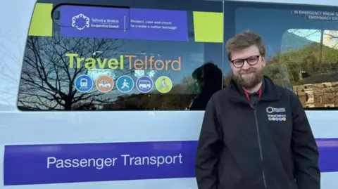 Telford and Wrekin Council Public transport officer Dylan Lawson is stood next to a minibus, which carries 'Travel Telford' and 'Passenger Transport' signage. He is wearing a black council-branded fleece jacket and black glasses.