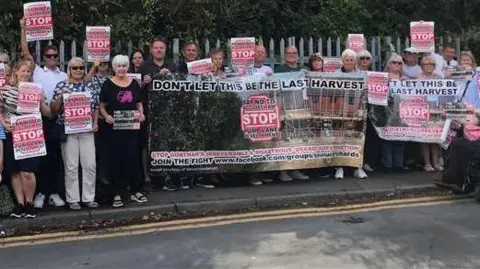 A group of people standing on a footpath holding signs and protesting against a proposed housing development. 