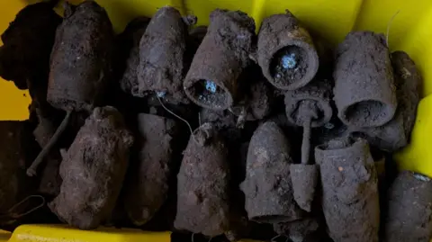 Mark Mather A pile of rusted and muddy bombs in a yellow box. They are shaped like bullets but larger.