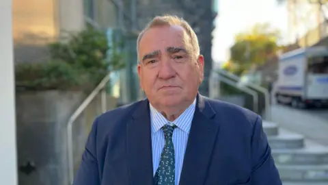 A man with a green tie and striped short wearing a blue suit looking towards camera. 