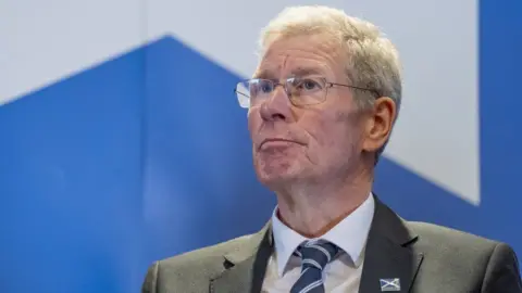 Kenny MacAskill, who has short grey hair, sits in front of a blue background. He is wearing a grey suit, white shirt and stripy tie, with a saltire badge on his jacket. 