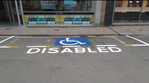 Government of Jersey A parking space with fresh painting of a disabled blue symbol with DISABLED painted in white below on the road, with a stone paved path behind and buildings.