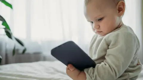 A baby in a grey jumper sits on a bed looking at a black phone.