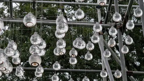 A frame with scores of clear round bottles full of water hanging on thread