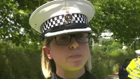 Supt Jo Grantham. She is a female police officer in a hat with a peak and a police badge. She has black, almost rectangular spectacles, made perhaps from plastic or acetate. Jo has blonde hair