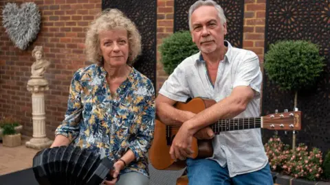 Catie Alison is holding a concertina and David is holding an acoustic guitar