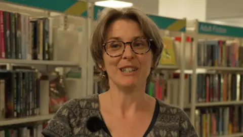 The image shows a woman wearing a patterned dress, with tortoiseshell glasses and light brown hair, standing in front of library bookshelves.