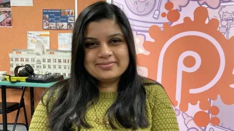 Hema Badger-Mistry has long dark hair and is smiling. She is wearing a lime green jumper.