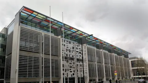 The photo shows a modern six-storey building, with a canopy of coloured glass panels extending from the roof.